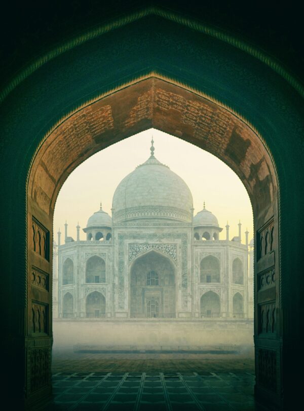 The iconic Taj Mahal viewed through a historic archway, enveloped in mist during sunrise in Agra, India.